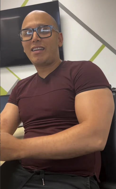 Bald man wearing glasses and maroon t-shirt smiling at camera with arms crossed indoors
